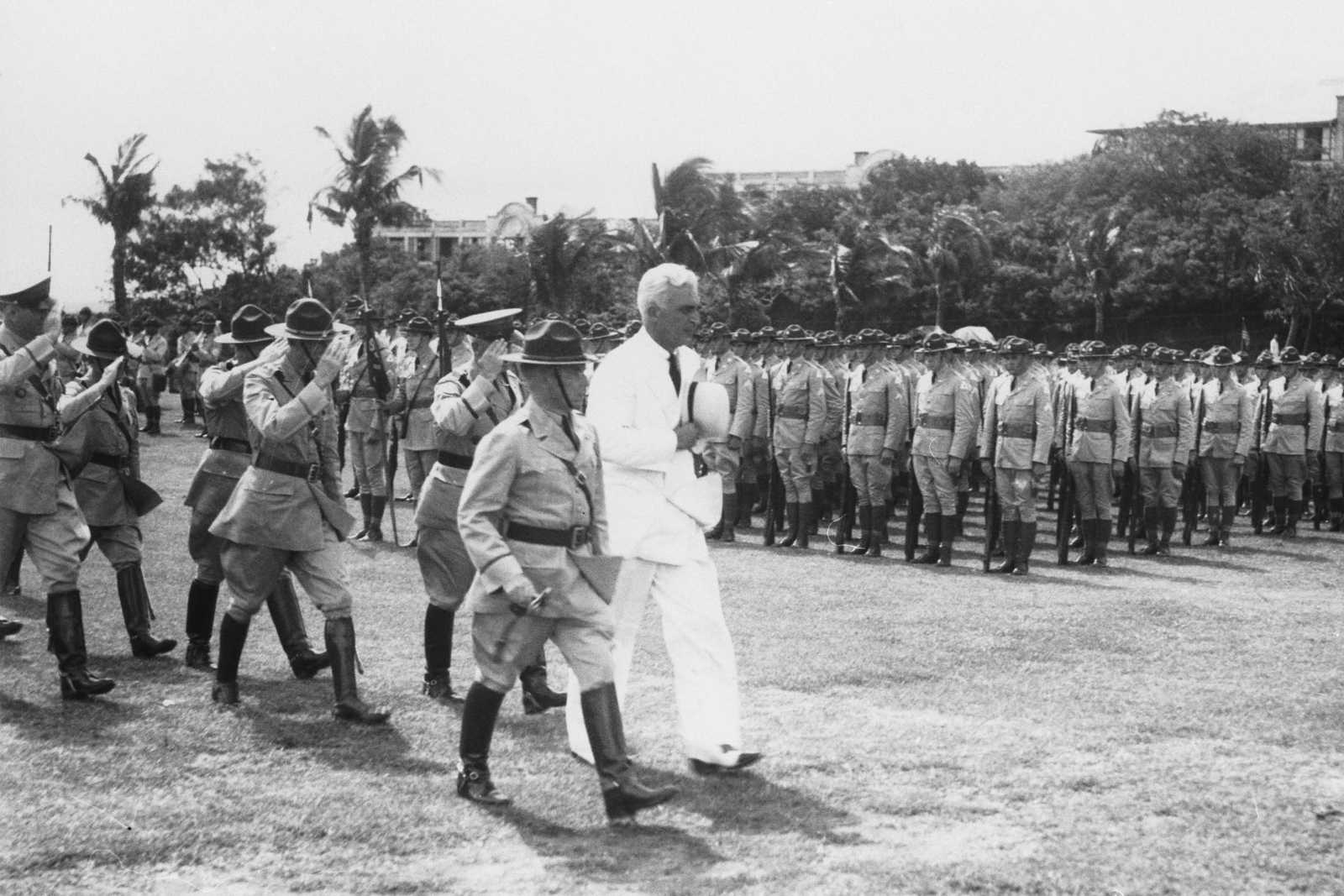 Filipino officers and American officer in white suit pass by Philippine soldiers in formation