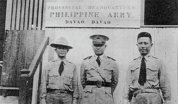 Black and white photo of three uniformed Filipino men standing under a sign that reads 