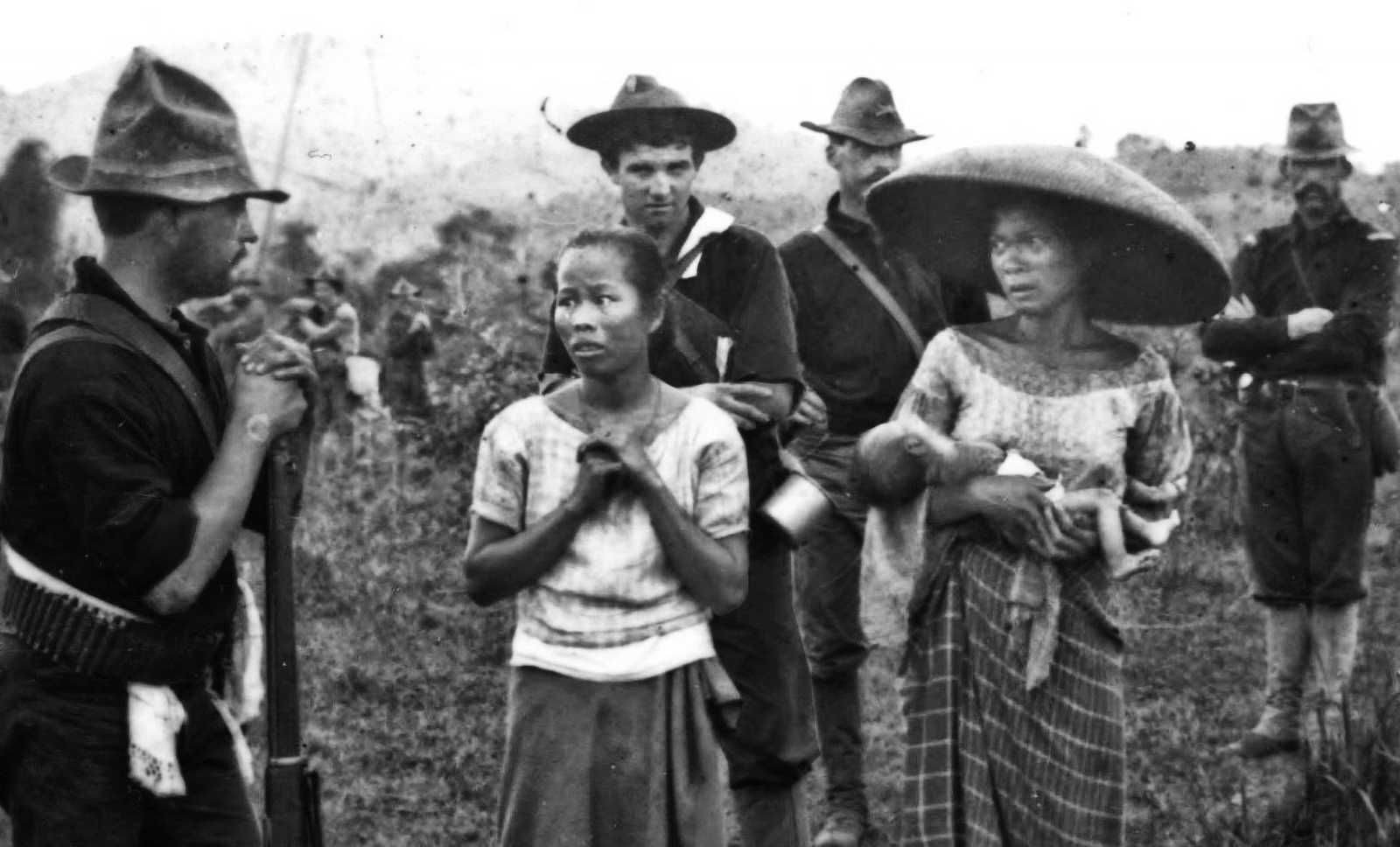 Black and white photo of Filipino women in a field (one holding a child) surrounded by American soldiers with rifles