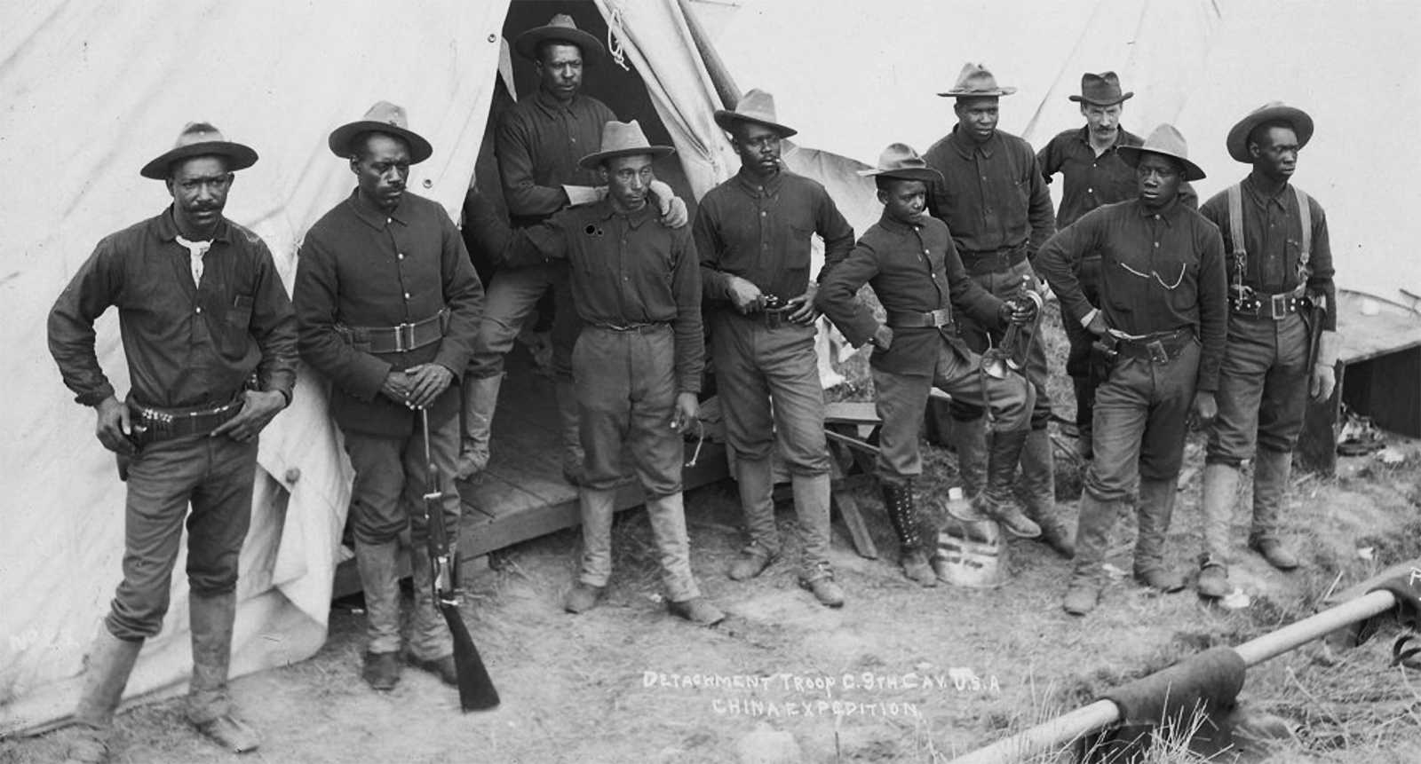 Black and white photo of African American soldiers standing outside their tent in uniform, some holding rifles