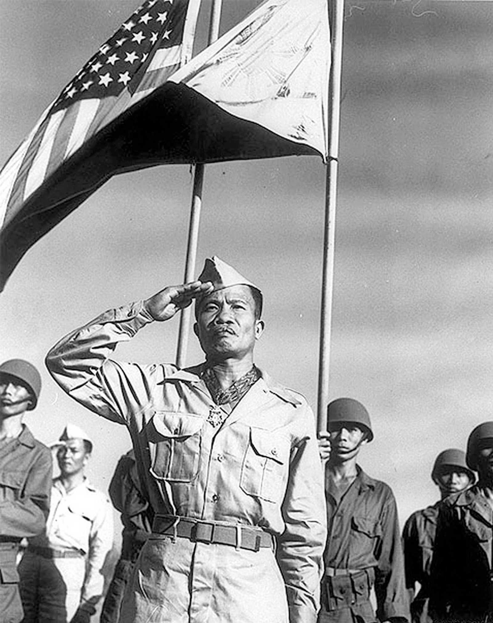 Photo of saluting Filipino soldier, Jose Calugas, standing under the American and Filipino flags with other soldiers standing at attention behind